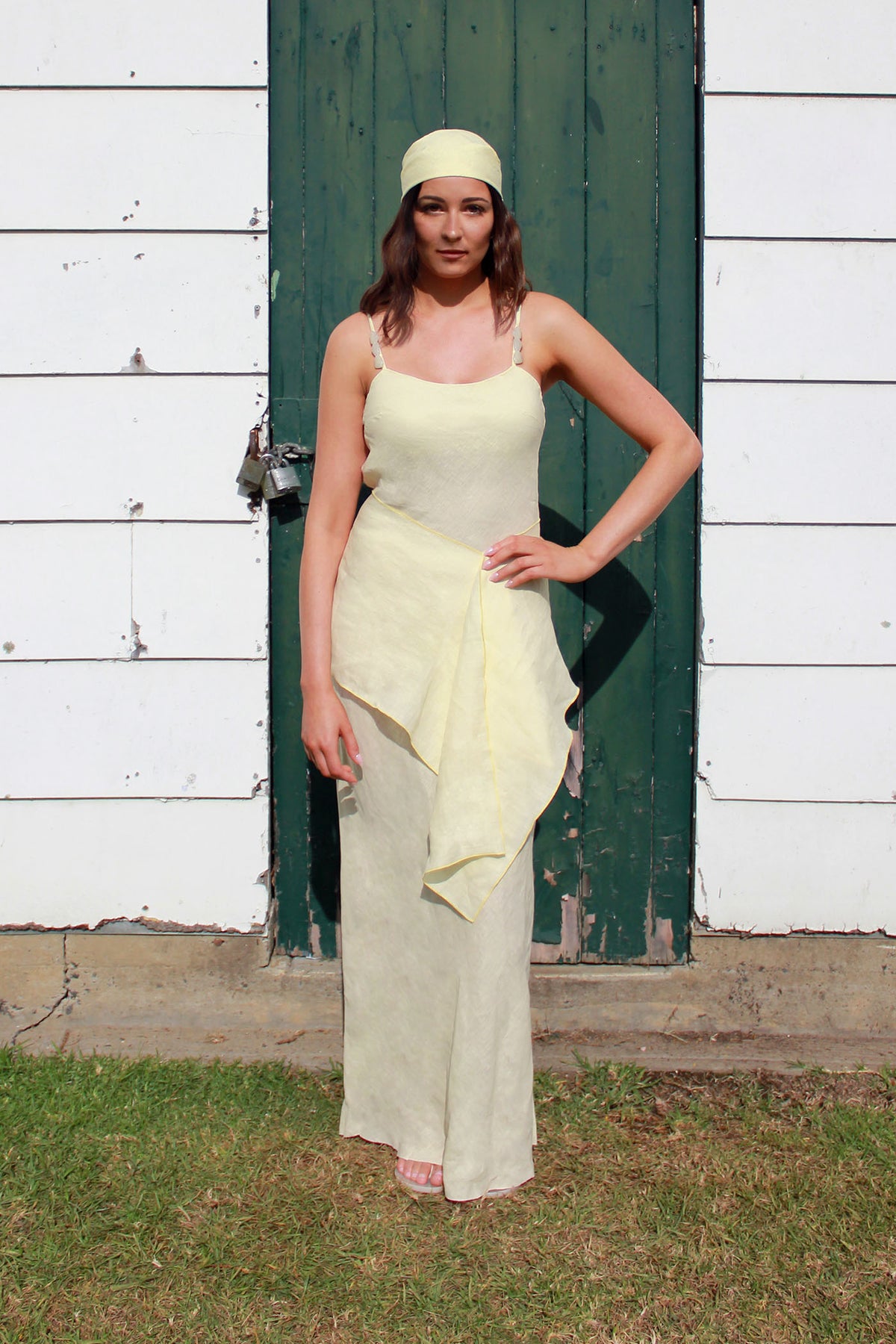A woman wearing a pale yellow sleeveless maxi dress with a draped wrap-style skirt and a matching headscarf, standing outdoors in front of a white wooden wall and a dark green door.