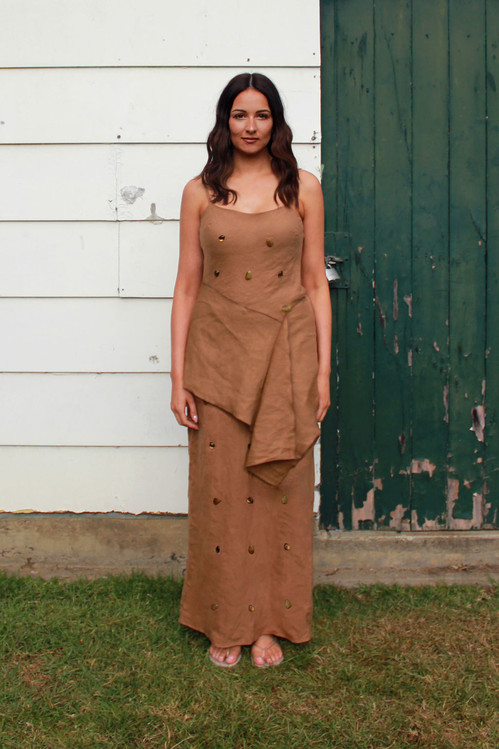 A woman wearing a brown sleeveless maxi dress with small gems and stones embellishments, standing outdoors on grass in front of a white wooden wall and a dark green distressed door.