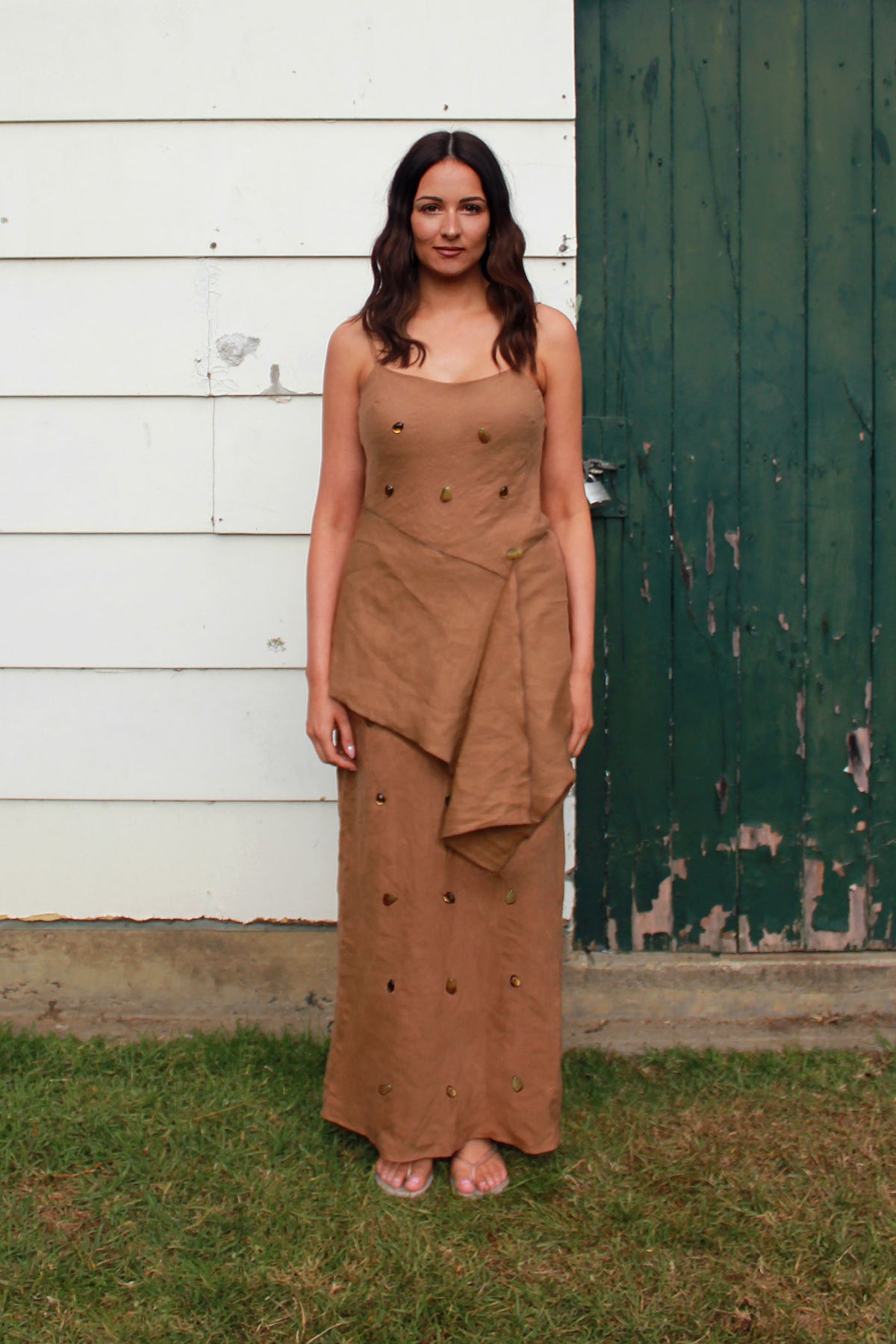 A woman wearing a brown sleeveless maxi dress with small gems and stones embellishments, standing outdoors on grass in front of a white wooden wall and a dark green distressed door.